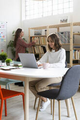 Businesswoman working on laptop with colleague searching book in background at office - GIOF09379