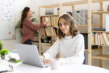 Young businesswoman using laptop with colleague standing in background at office - GIOF09381