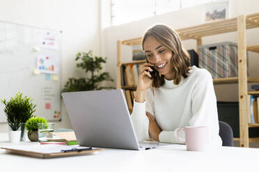 Lächelnde Geschäftsfrau, die mit ihrem Handy telefoniert, während sie im Büro am Laptop arbeitet - GIOF09382