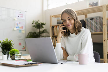 Businesswoman talking on mobile phone while working on laptop at office - GIOF09383