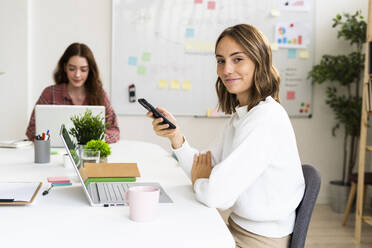 Lächelnde Geschäftsfrau, die ein Mobiltelefon benutzt, während ihr Kollege im Hintergrund an einem Laptop arbeitet - GIOF09387