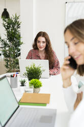 Lächelnde junge Frau, die an einem Laptop arbeitet, mit einer Geschäftsfrau, die mit einem Mobiltelefon im Vordergrund im Büro spricht - GIOF09389