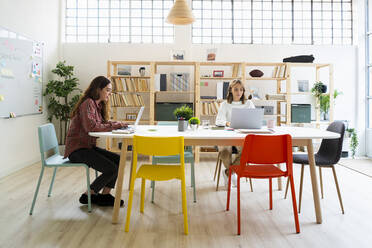 Businesswoman and colleague working on laptop while sitting by table at office - GIOF09390