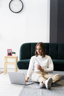 Geschäftsfrau mit Kaffeetasse und Laptop, während sie im Büro auf dem Boden sitzt - GIOF09401