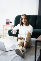 Lächelnde Geschäftsfrau mit Kaffeetasse, die ein Mobiltelefon benutzt, während sie auf dem Boden im Büro sitzt - GIOF09402