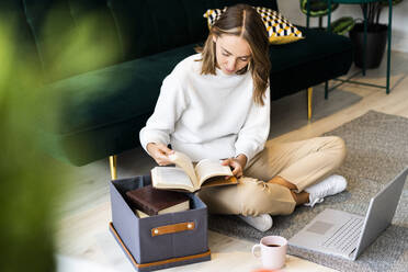 Businesswoman reading book while sitting by laptop and box on floor at office - GIOF09407