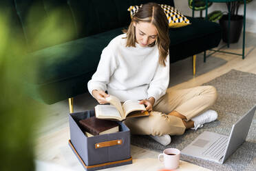 Geschäftsfrau, die ein Buch liest, während sie neben einem Laptop und einer Kiste auf dem Boden im Büro sitzt - GIOF09407