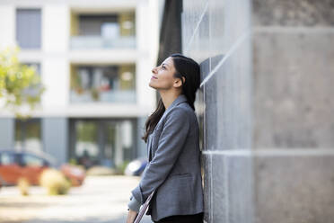 Businesswoman with laptop looking up while leaning on wall - SGF02717