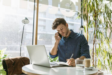 Geschäftsmann schreibt in Tagebuch, während er am Tisch in einem Café mit seinem Mobiltelefon spricht - VABF03745