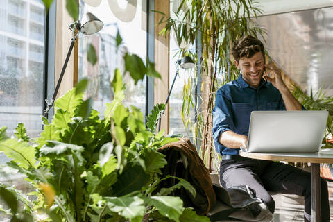 Smiling male freelancer talking on mobile phone while using laptop at table in cafe stock photo