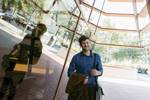 Smiling young businessman with blazer and bag standing outside cafe stock photo