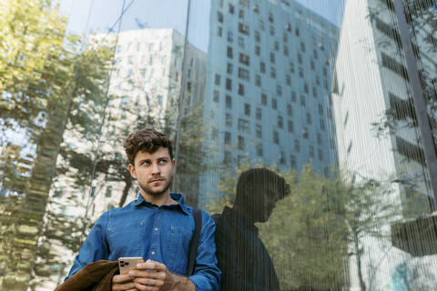 Nachdenklicher junger Geschäftsmann lehnt an der Glaswand eines modernen Bürogebäudes, lizenzfreies Stockfoto