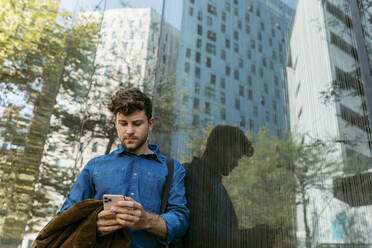 Handsome young businessman using smart phone while leaning on glass wall of modern office building - VABF03769