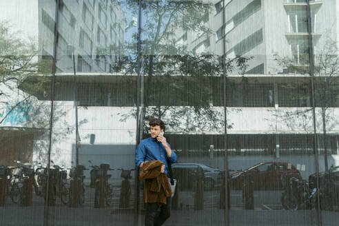 Junger Geschäftsmann, der sich an die Wand eines modernen Glasgebäudes lehnt, während er mit seinem Smartphone telefoniert - VABF03770