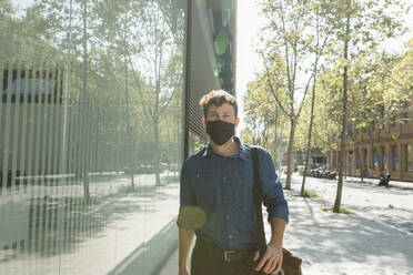 Young businessman wearing face mask while walking on footpath in city during coronavirus crisis - VABF03773