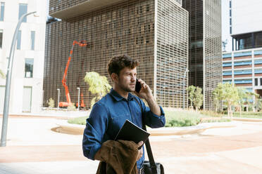 Handsome young businessman talking on smart phone while walking on footpath in city - VABF03777