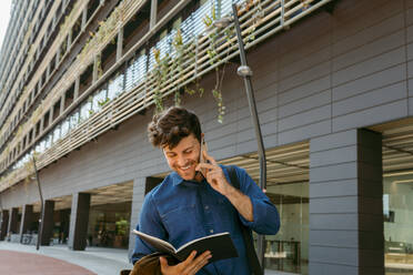 Smiling young businessman looking at diary while talking on smart phone outside office building - VABF03778