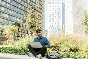 Geschäftsmann schaut weg, während er mit seinem Laptop auf einer Stützmauer vor einem Bürogebäude sitzt - VABF03779