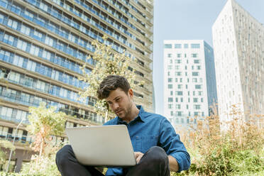 Gut aussehender Geschäftsmann, der einen Laptop benutzt, während er vor einem modernen Gebäude sitzt - VABF03780