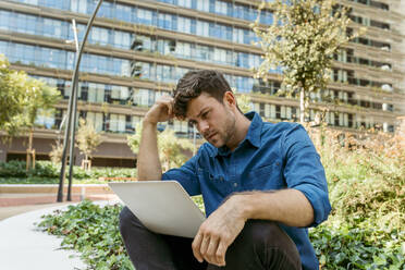 Confused young businessman using laptop while sitting against office buildings - VABF03782