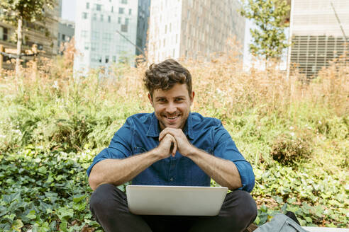 Lächelnder junger Geschäftsmann sitzt mit Laptop vor Pflanzen - VABF03785
