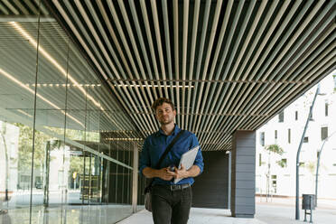Young businessman walking with laptop on walkway at modern office building - VABF03787
