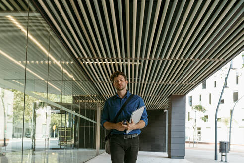 Junger Geschäftsmann geht mit Laptop auf dem Gehweg eines modernen Bürogebäudes - VABF03787