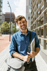 Smiling young businessman standing on footpath in city during sunny day - VABF03792