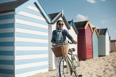 Frau zu Fuß mit Fahrrad auf Sand durch Strandhütten am sonnigen Tag - AJOF00448