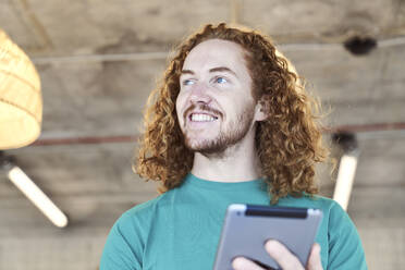 Männlicher Hipster mit langen Haaren, der wegschaut, während er ein digitales Tablet zu Hause benutzt - FMKF06643