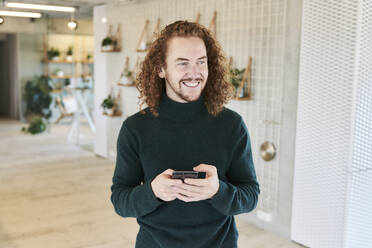 Cheerful mid adult man looking away while using mobile phone at studio apartment - FMKF06648