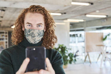 Mann mit Gesichtsschutzmaske fotografiert mit dem Handy in einer Studiowohnung - FMKF06651