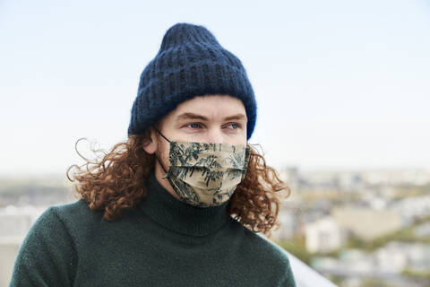 Thoughtful man with curly long hair wearing protective face mask and beanie on rooftop stock photo