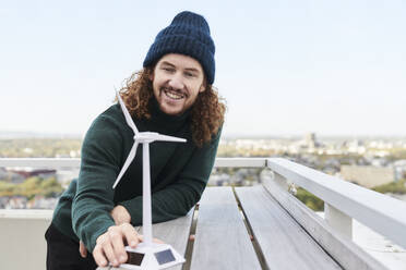 Lächelnder männlicher Hipster mit Windmühlenmodell auf einer Gebäudeterrasse gegen den Himmel - FMKF06653