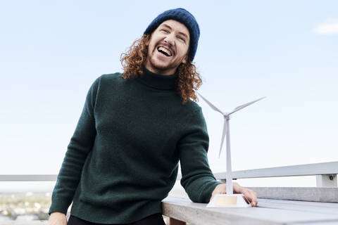 Cheerful man with windmill model on building terrace against sky stock photo