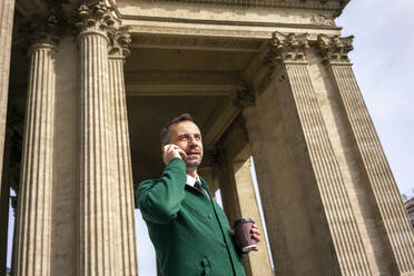 Geschäftsmann mit Kaffeetasse, der mit seinem Handy telefoniert, während er vor den architektonischen Säulen der Kasaner Kathedrale in Sankt Petersburg, Russland, steht - VPIF03179