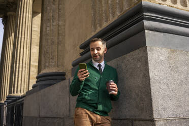 Businessman with coffee cup using mobile phone while leaning on pillar of Kazan Cathedral at Saint Petersburg, Russia - VPIF03180