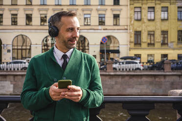 Businessman with headphones looking away while using mobile phone in city - VPIF03184