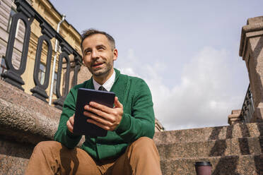 Lächelnder Geschäftsmann, der ein digitales Tablet benutzt, während er auf einer Treppe in der Stadt sitzt - VPIF03187