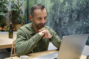 Lächelnder Geschäftsmann mit dem Kopf in den Händen bei der Arbeit am Laptop, während er im Café sitzt - VPIF03192