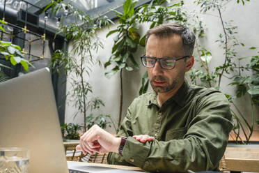 Geschäftsmann mit Brille prüft die Zeit, während er in einem Café am Laptop arbeitet - VPIF03193
