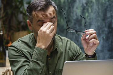 Müder Geschäftsmann mit dem Kopf in den Händen bei der Arbeit am Laptop, während er im Café sitzt - VPIF03200