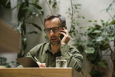 Businessman wearing eyeglasses writing in book while working at cafe - VPIF03209