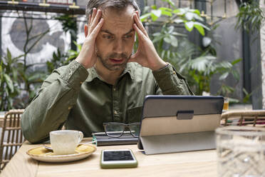 Geschäftsmann mit Kopf in den Händen, der ein digitales Tablet benutzt, während er in einem Café sitzt - VPIF03217