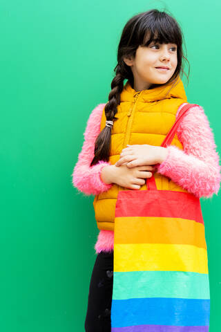 Girl carrying colorful bag looking away while standing against green wall stock photo