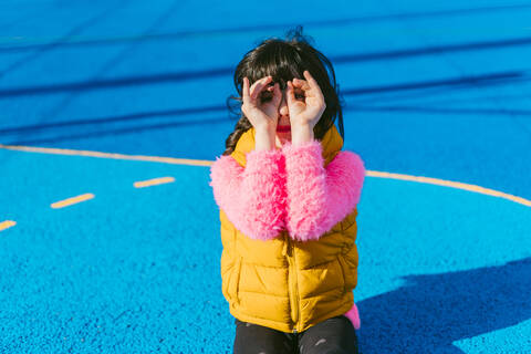 Girl forming binoculars with fingers while sitting at sports court stock photo