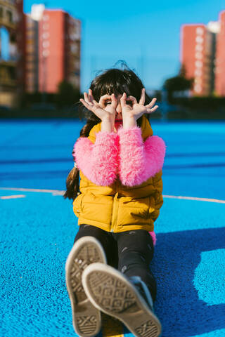 Girl forming binoculars with fingers while sitting on sports court during sunny day stock photo