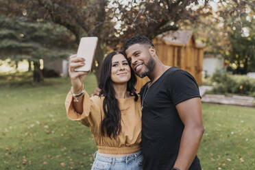 Smiling woman taking selfie with man through mobile phone while standing at backyard - SMSF00389
