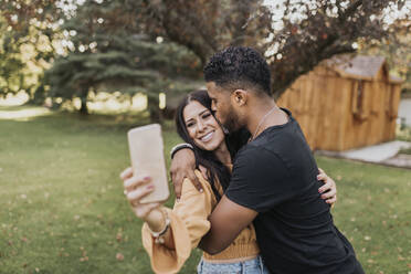 Mann küsst Frau, die ein Selfie mit ihrem Mobiltelefon macht, während sie im Hinterhof steht - SMSF00390