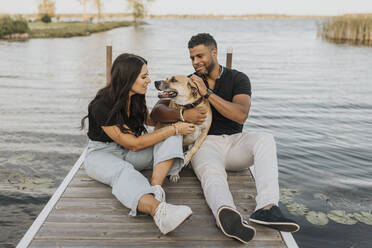 Paar spielt mit Hund und sitzt auf einem Pier am See - SMSF00425